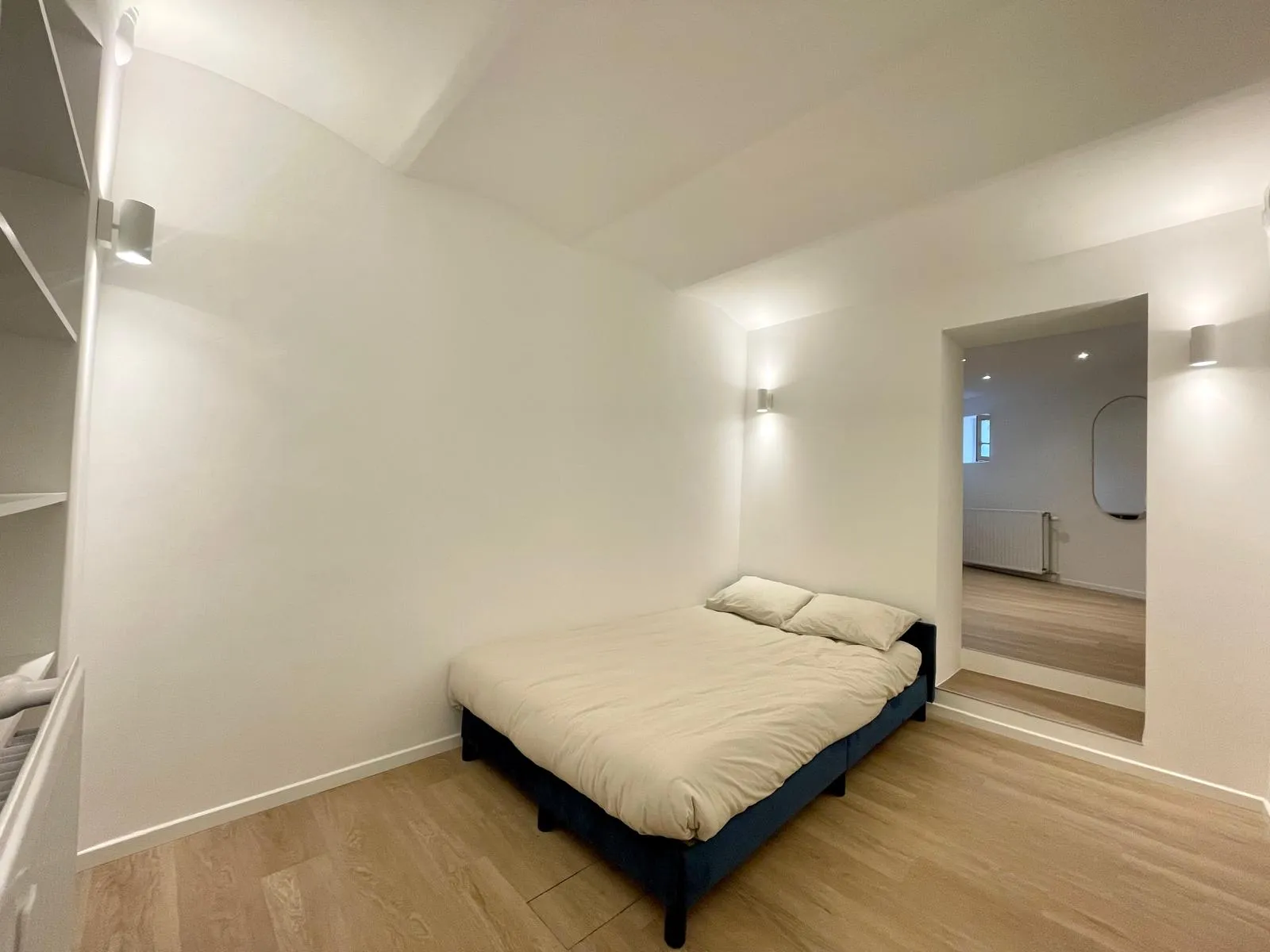 Chambre aux murs blancs et appliques murales, étagères encastrées, passage vers la salle d'eau