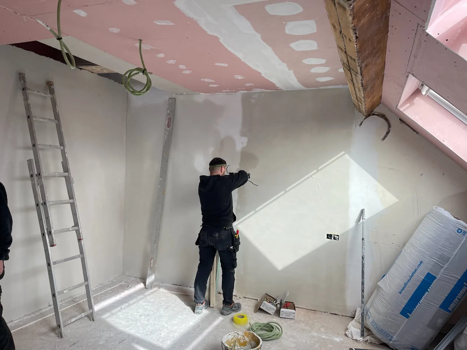 Plafonneur lissant un mur à l'enduit sous une charpente en cours d'isolation