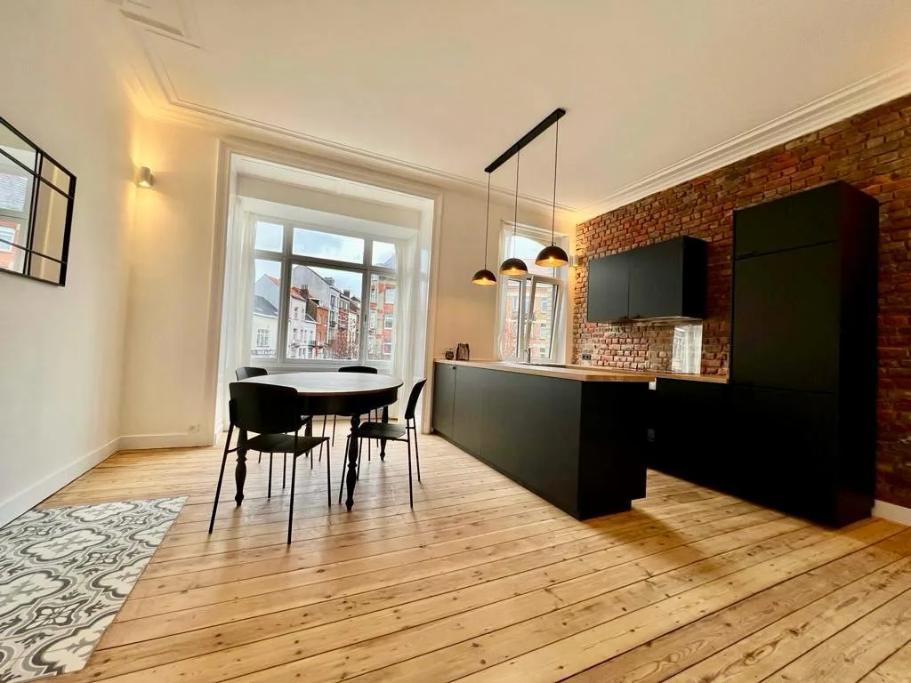 Salle à manger avec cuisine noire sur mur de briques apparentes, bow-window à moulures et tapis de carreaux de ciment